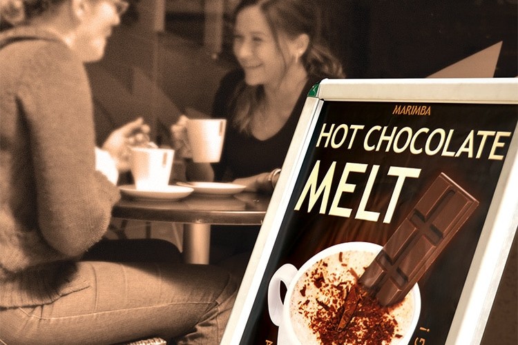 Promotion Design poster signage for Marimba's Ice Chocolate Melt in front of two people enjoying a cup of hot chocolate